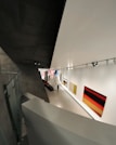 A modern art gallery with a sleek, angular architectural design. The interior features large, brightly colored abstract paintings on the walls. A solitary figure walks through the exhibition space, which is illuminated by overhead track lighting, highlighting the artwork on display.
