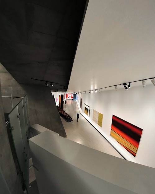 A modern art gallery with a sleek, angular architectural design. The interior features large, brightly colored abstract paintings on the walls. A solitary figure walks through the exhibition space, which is illuminated by overhead track lighting, highlighting the artwork on display.
