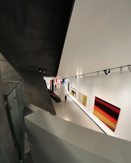 A modern art gallery with a sleek, angular architectural design. The interior features large, brightly colored abstract paintings on the walls. A solitary figure walks through the exhibition space, which is illuminated by overhead track lighting, highlighting the artwork on display.