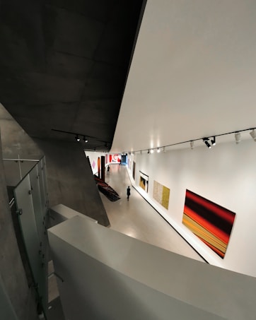 A modern art gallery with a sleek, angular architectural design. The interior features large, brightly colored abstract paintings on the walls. A solitary figure walks through the exhibition space, which is illuminated by overhead track lighting, highlighting the artwork on display.