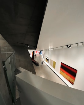 A modern art gallery with a sleek, angular architectural design. The interior features large, brightly colored abstract paintings on the walls. A solitary figure walks through the exhibition space, which is illuminated by overhead track lighting, highlighting the artwork on display.
