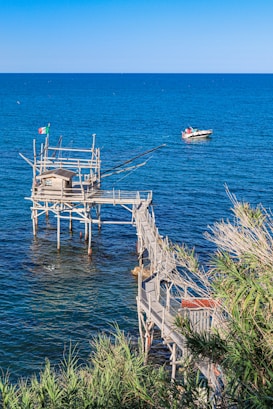 A wooden fishing platform extends over a clear blue sea, connected by a narrow walkway. A small boat floats nearby, and green foliage surrounds the area. An Italian flag is visible on the structure.