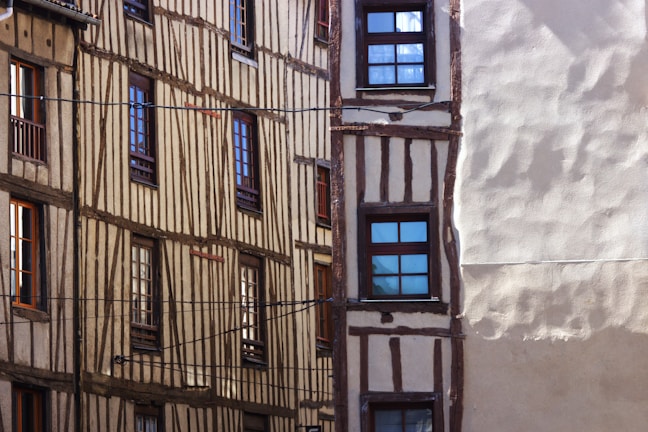 An architectural view of a historic building featuring half-timbered walls with vertical and horizontal wooden frames. Several windows with dark frames are visible. Sunlight casts shadows onto the facade, enhancing the textures.