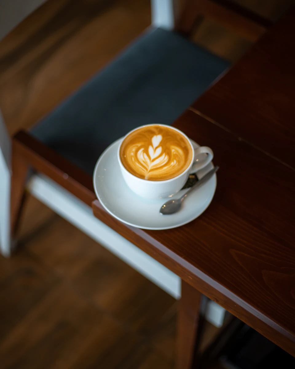 The image shows a white coffee cup with an intricate latte art design on its surface, placed on a wooden table. The cup is accompanied by a silver spoon and saucer, suggesting it's ready to be enjoyed. In the background, there are two chairs - one black and one gray - positioned against the wall.