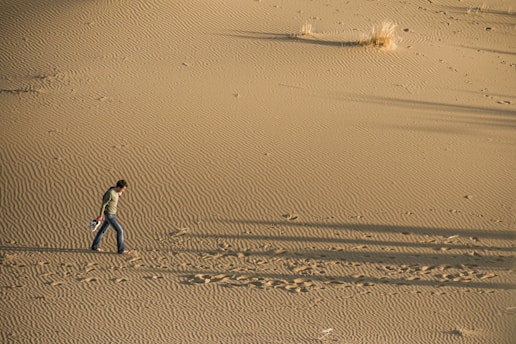 A person walks barefoot across a vast expanse of rippled sand dunes. The individual carries shoes in one hand and leaves footprints behind. Sparse patches of dry grass are visible in the background, casting long shadows in the warm, golden light.