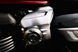 The image features a close-up view of a metallic component, likely part of an engine or machinery. The metal has a shiny, brushed finish with visible screws and grooves. The background includes other parts with different textures and a hint of red, possibly indicating a vehicle or machine housing.