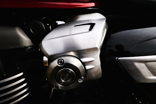 The image features a close-up view of a metallic component, likely part of an engine or machinery. The metal has a shiny, brushed finish with visible screws and grooves. The background includes other parts with different textures and a hint of red, possibly indicating a vehicle or machine housing.