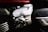 The image features a close-up view of a metallic component, likely part of an engine or machinery. The metal has a shiny, brushed finish with visible screws and grooves. The background includes other parts with different textures and a hint of red, possibly indicating a vehicle or machine housing.