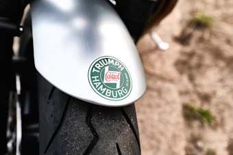Close-up of a motorcycle tire and fender featuring a round green and white logo with the words 'Triumph Hamburg' and a Castrol emblem. The image is taken from an angle showing the tread pattern of the front tire.