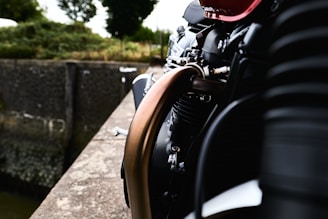 A close-up view of a motorcycle engine with a vivid red tank and metallic components standing out against a blurred background of greenery and a stone wall. The image focuses on the intricate details of the engine pipes and components.
