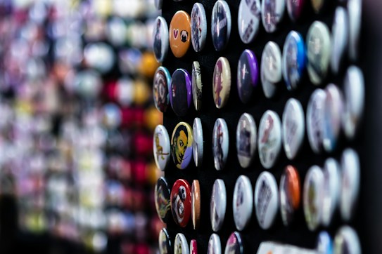 A collection of colorful round badges displayed in rows on a dark surface. Each badge has a unique design or image, ranging from patterns to illustrations. The background is softly blurred, emphasizing the sharp details of the badges in the foreground.
