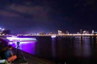 A nighttime cityscape with illuminated buildings along a river. Lights from the buildings and a brightly lit boat reflect on the calm water. In the foreground, a person sitting beside a tripod with a camera is visible, overlooking the river.