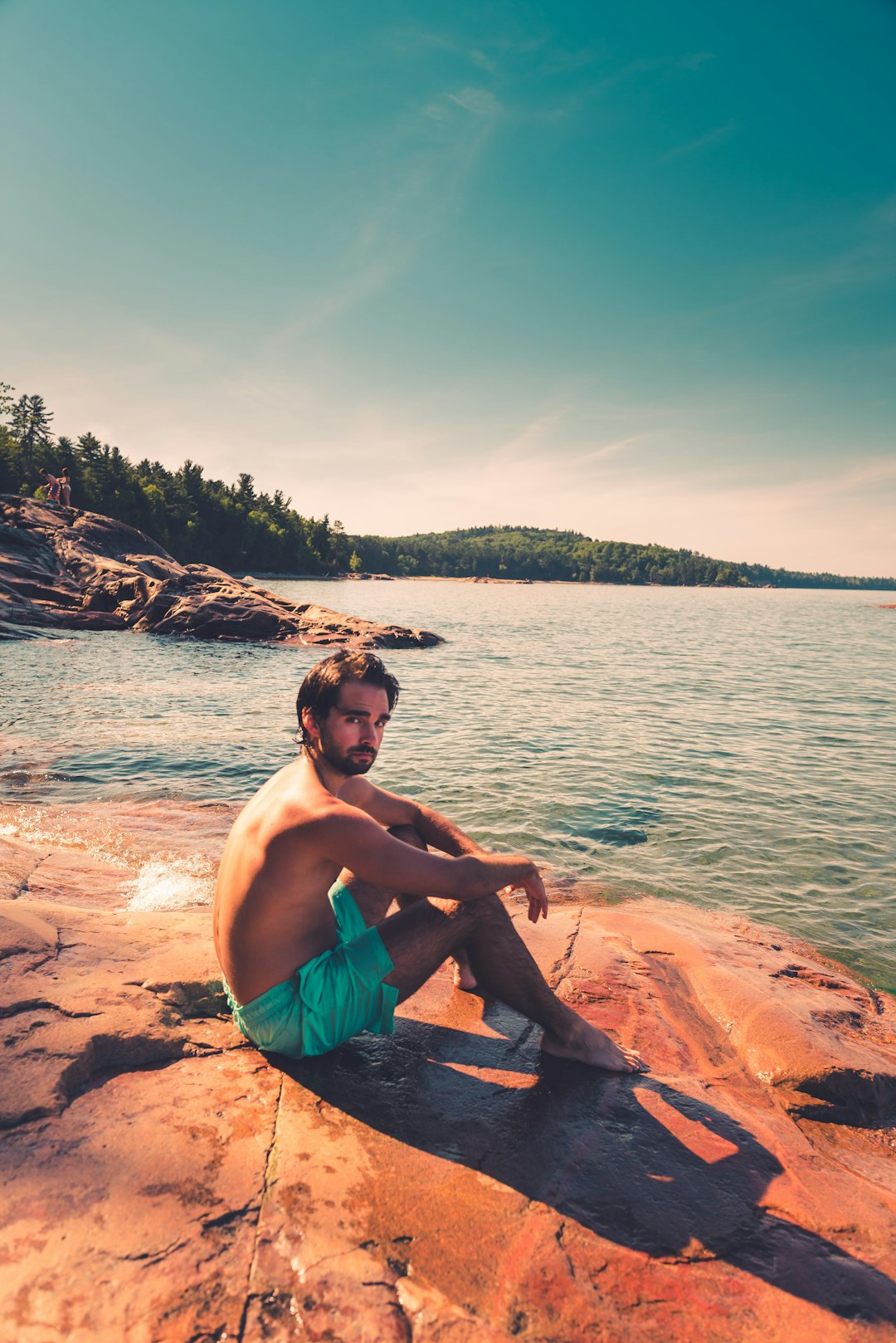 topless man in green shorts sitting on brown rock near body of water during daytime