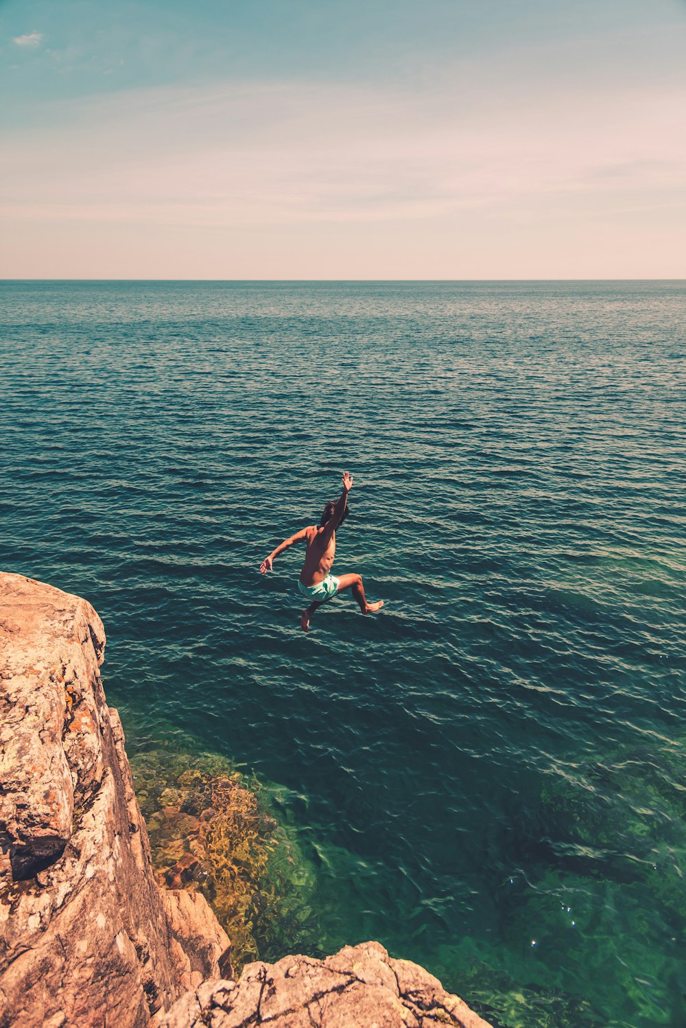 woman in blue shorts jumping on brown rock formation near body of water during daytime