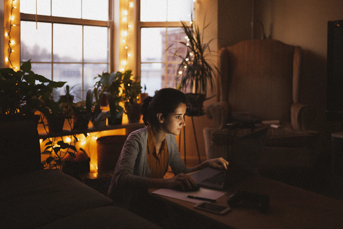 A woman works on a laptop at a table by the window in the evening, lit by string lights and houseplants