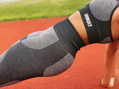 A person is performing a plank exercise on a red track field. They are wearing a matching athletic outfit consisting of a grey and black sports bra and leggings, with the word 'UNRULY' printed on the top. The background includes a blurred view of green grass.