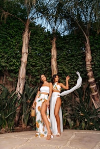 Two women stand outdoors against a lush background of tall palm trees and dense greenery. They are dressed in elegant beachwear, with one wearing a white tube top and a pineapple-patterned skirt, and the other in a matching set with a flowing sheer cover-up. The setting appears tropical and serene.