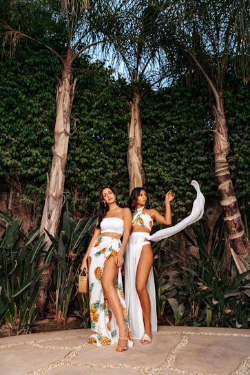 Two women stand outdoors against a lush background of tall palm trees and dense greenery. They are dressed in elegant beachwear, with one wearing a white tube top and a pineapple-patterned skirt, and the other in a matching set with a flowing sheer cover-up. The setting appears tropical and serene.