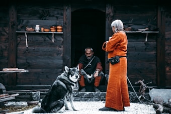 An elderly person wearing an orange robe stands facing a man seated in a dark entrance. A large dog sits nearby, looking towards the viewer. The setting is rustic, with a wooden structure in the background and shelves holding clay pots.
