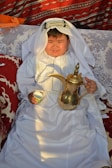 A child dressed in traditional Middle Eastern attire sits on a patterned couch. The child is wearing a white robe with intricate embroidery and a headscarf, and holds a traditional Arabic coffee pot and a small cup. The background features vibrant, patterned textiles with shades of red and white.