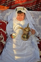 A child dressed in traditional Middle Eastern attire sits on a patterned couch. The child is wearing a white robe with intricate embroidery and a headscarf, and holds a traditional Arabic coffee pot and a small cup. The background features vibrant, patterned textiles with shades of red and white.