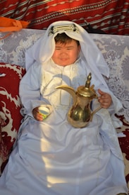 A child dressed in traditional Middle Eastern attire sits on a patterned couch. The child is wearing a white robe with intricate embroidery and a headscarf, and holds a traditional Arabic coffee pot and a small cup. The background features vibrant, patterned textiles with shades of red and white.