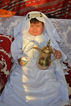 A child dressed in traditional Middle Eastern attire sits on a patterned couch. The child is wearing a white robe with intricate embroidery and a headscarf, and holds a traditional Arabic coffee pot and a small cup. The background features vibrant, patterned textiles with shades of red and white.