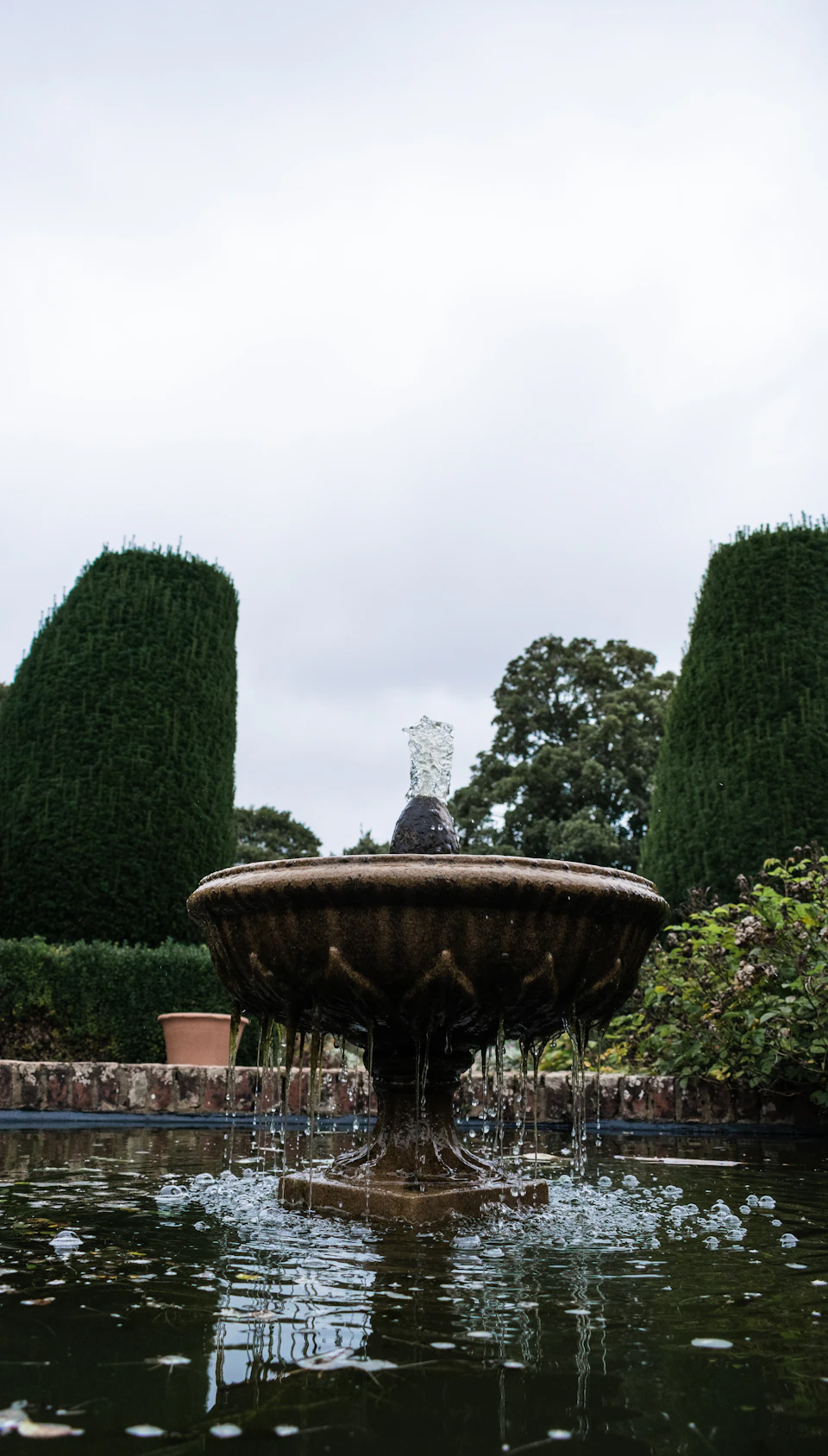 The image depicts a serene garden scene featuring a large stone fountain with water cascading down its center, surrounded by lush green trees and bushes. The sky above is overcast, casting a soft light on the tranquil setting.