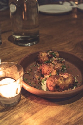 A warmly lit dining table featuring a brown ceramic plate with several golden-brown croquettes garnished with herbs and diced onions. A small glass candle holder with a lit candle adds a cozy ambiance to the scene. The background displays blurred elements including a glass bottle and a white plate.