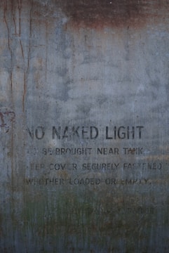 A weathered and rusty metal surface with faded text that reads 'NO NAKED LIGHT IS BE BROUGHT NEAR TANK KEEP COVER SECURELY FASTENED WHETHER LOADED OR EMPTY.' The text is partially obscured by rust and grime.
