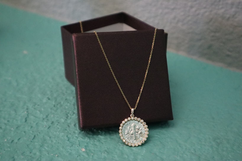 A gold chain necklace with a circular pendant that features a central teal-colored design encircled by clear stones. The necklace is draped over a dark brown jewelry box, both of which rest on a light teal surface. The background is a textured light blue-gray wall.