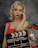 A woman with blonde hair and red lipstick is wearing a red dress and holding a film clapperboard. There is a disco ball visible in the background.