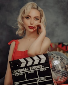 A woman with blonde hair and red lipstick is wearing a red dress and holding a film clapperboard. There is a disco ball visible in the background.