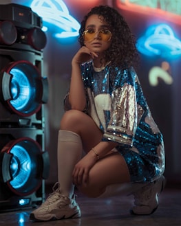 A person with curly hair wearing yellow-tinted glasses and a sequined blue and silver oversized shirt is crouching next to a large speaker with glowing blue lights. The background features neon light designs.