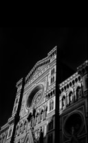 A dramatic black and white image of an ornate, gothic-style cathedral facade featuring intricate carvings, arches, and a large circular rose window. The lighting emphasizes the detailed architectural features, casting deep shadows that add depth and texture.