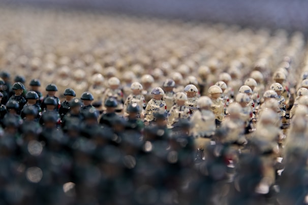 A densely packed assembly of tiny toy army figurines, arranged in rows. The figures are detailed with distinct camouflage patterns, predominantly in shades of green and beige, giving the illusion of a military formation. The focus shifts from blurry, indistinct figures in the foreground to sharply defined ones in the background, creating a depth of field effect.