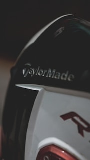 A close-up view of a golf club head with the brand name TaylorMade prominently displayed. The design features a combination of dark and light colors with a hint of red detail.