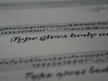 A close-up view of typeface samples printed on a sheet of paper. The text is in different styles and fonts, highlighting decorative and serif characteristics. The paper appears slightly angled, emphasizing the artistic nature of typography.