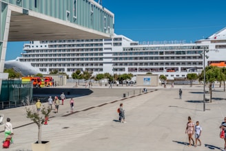 A large cruise ship is docked at a port, with people walking along a plaza nearby. There are several trees and a building with the sign 'VILLA MEDIT' visible. A fire truck is parked close to the docked ship, and the weather appears to be sunny with clear blue skies.