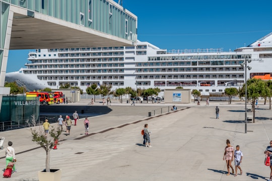 A large cruise ship is docked at a port, with people walking along a plaza nearby. There are several trees and a building with the sign 'VILLA MEDIT' visible. A fire truck is parked close to the docked ship, and the weather appears to be sunny with clear blue skies.
