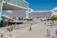 A large cruise ship is docked at a port, with people walking along a plaza nearby. There are several trees and a building with the sign 'VILLA MEDIT' visible. A fire truck is parked close to the docked ship, and the weather appears to be sunny with clear blue skies.