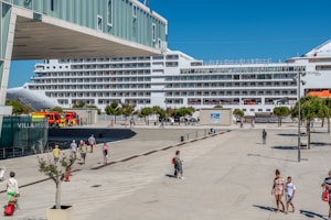 A large cruise ship is docked at a port, with people walking along a plaza nearby. There are several trees and a building with the sign 'VILLA MEDIT' visible. A fire truck is parked close to the docked ship, and the weather appears to be sunny with clear blue skies.