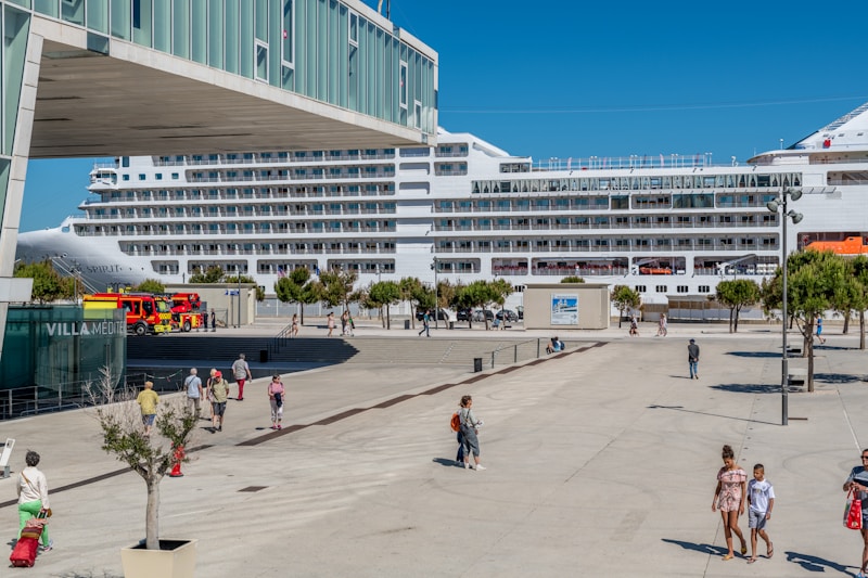 A large cruise ship is docked at a port, with people walking along a plaza nearby. There are several trees and a building with the sign 'VILLA MEDIT' visible. A fire truck is parked close to the docked ship, and the weather appears to be sunny with clear blue skies.
