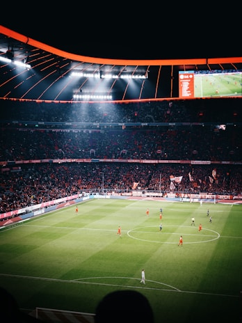 A large football stadium filled with spectators watching a game in progress. The stadium is brightly lit, with players in red and dark uniforms scattered across the green field. The scoreboard is visible, displaying game information. The atmosphere appears lively and energetic, with a sea of fans in the stands.