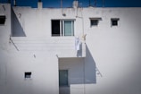 A white building exterior with several small windows. A single white towel is hanging on a clothesline on the balcony, casting a shadow against the wall. The architecture appears minimalistic with a clean and uncluttered facade.