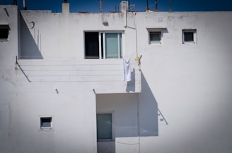 A white building exterior with several small windows. A single white towel is hanging on a clothesline on the balcony, casting a shadow against the wall. The architecture appears minimalistic with a clean and uncluttered facade.