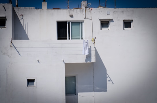 A white building exterior with several small windows. A single white towel is hanging on a clothesline on the balcony, casting a shadow against the wall. The architecture appears minimalistic with a clean and uncluttered facade.