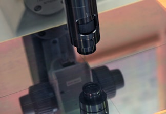 A close-up of a microscope lens focusing on a reflective surface. The equipment is primarily metallic with a dark finish, and there is a soft reflection visible on the surface below it. The background shows machinery parts, blurred to focus on the microscope.