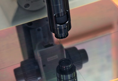 A close-up of a microscope lens focusing on a reflective surface. The equipment is primarily metallic with a dark finish, and there is a soft reflection visible on the surface below it. The background shows machinery parts, blurred to focus on the microscope.