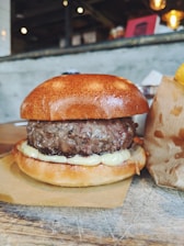 A juicy hamburger with a thick patty sitting on a slightly toasted bun. The burger features melted cheese beneath the patty, with a backdrop of a rustic interior with blurred lights and a cardboard container on the side.